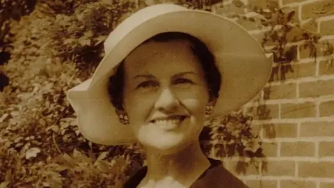 McKay family Muriel McKay photographed in a black and white sepia picture. She is wearing a cream summer hat and a dark jacket open at the neck, and cluster earrings. She is smiling at the camera and standing in front of a brick wall with a trailing plant like wisteria growing in the background.