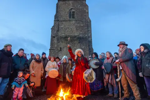 Getty Images People gather on Glastonbury Tor as they take part in a winter solstice ceremony. The church on the hill can be seen in the background, with people standing around a woman dressed in red holding a drum and a bonfire.