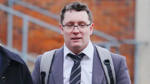 Ben Birchall/PA Wire Christopher Trybus arriving outside court wearing a white collared shirt, striped tie and grey blazer. He has dark grey hair and is wearing rectangular framed glasses and a grey rucksack. 