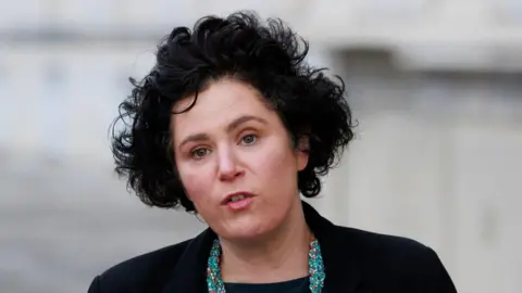 PA Ms Hanna with short black curly hair speaks to the camera. She is wearing make-up, a black blazer and black top with a blue beaded necklace. The background is blurred.