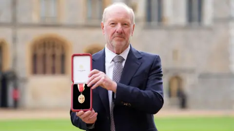 PA Media Sir Alan Bates holds his medal in a red case after he is made a knight at Windsor Castle. Sir Bates is balding with grey hair and has a short beard. He is wearing a navy suit, with a white collared tie and a blue and brown circular tie. He is looking just beyond the camera with a neutral expression on his face.