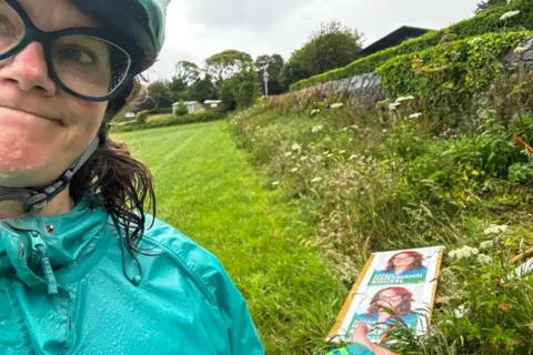 Sarah Hansmann-Rouxel A woman with wet, shoulder length brown wavy hair looking at the camera with her lips pushed together. She is wearing a cycle helmet and large glasses. She is pointing to some of her vandalised election posters in a field behind her.