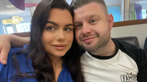 Josh Bashford wears a black and white t-shirt. He has short brown hair and a cropped beard. He hugs his wife Chloe who has long dark hair and wears a bright blue blouse. Both of them are smiling. 