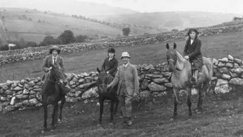 A black-and-white photograph showing three females on horseback and a man standing beside the middle horse in the 1920s. The man is dressed in a suit and hat, and the riders, two women and a younger girl, also wear traditional attire of the time, including jackets and hats. They are in hilly, open countryside bordered by dry stone walls.