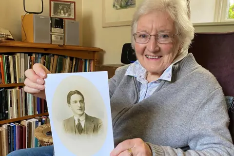 Libby MacRae in an armchair holding a photo of her uncle Donnie MacRae. She is in a living room at her home with bookshelves behind her chair. She is smiling at the camera. She has grey hair and glasses and is wearing a comfortable woollen zip sweater with a shirt underneath and blue jeans. 