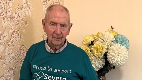 A man with short grey hair and a teal jumper a room with a large bunch of white flowers behind him