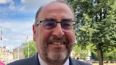 Image of a man with glasses and a stubble beard, smiling at the camera. In the background can be seen a city park, trees, a number of flags and large Georgian style buildings.