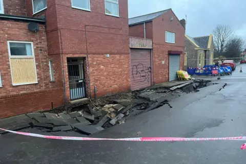 The sinkhole in front of a two-storey red bricked house. The grey tarmac road is splintered a caved in.