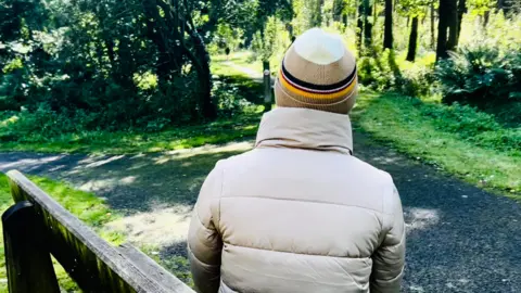 An anonymous photo of Kate taken from behind. She is sitting on a park bench and wearing a puffy cream-coloured jacket and a brown and white woolly hat with red,  white, blue and yellow bands. In the background are grass and trees.