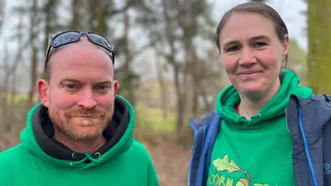 Alex Osborne and Michelle Sinsheimer looking directly at the camera. Both are wearing a green hoodie. Alex, on the left, has sunglasses on his head. He has a ginger beard with white patches. Michelle, on the right, is taller. She has dark hair and is wearing her hoodie under a blue jacket.