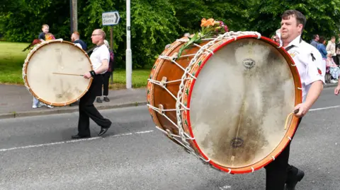Pacemaker Two band members playing Lambeg drums during a 12th July parade in Carrickfergus last year. The huge, wooden, double-sided drums are worn on straps across the front of their bodies and they stretch in length from their face to their knees. The drummers are both wearing white shirts and black trousers.