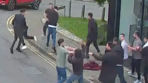 Image depicts multiple men fighting outside on the street. There are over ten men with some in the middle of their fight and others overlooking the ensuing brawl. 