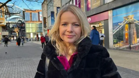 A woman, Susannah Streeter pictured on a high street. She has blonde hair and is wearing a pink top under a black fur coat. 