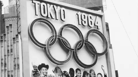 Getty Images A large sign displays the text “Tokyo 1964” above five interlocking Olympic rings. The structure appears outdoors, with tall buildings in the background and images of children under the sign.
