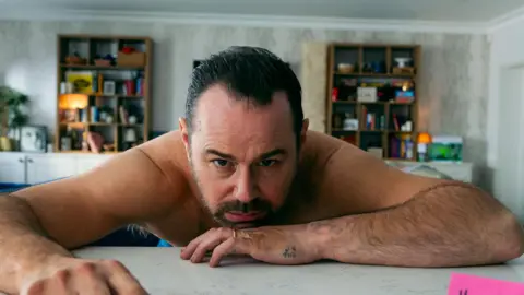 Danny Dyer is slumped on a white table, chin on his elbow and looking up at the camera. He has a dark beard and moustache and wears a gold wedding ring and no shirt. There is a small tattoo on the side of his hand. In the background are two bookshelves, one either side of him. His expression suggests he is feeling rough.