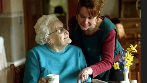 Getty/Matt Cardy File image of a carer leaning over the shoulder of an elderly lady who is sitting down and looking up at her. The elderly woman is wearing a blue jumper and has a cup in her right hand. The female care worker is wearing a dark green tabard over a burgundy coloured jumper.