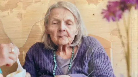 An elderly woman wearing purple, being fed yoghurt in a care home setting