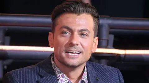 Paul Danan wearing a blue pinstripe blazer over a pink flowery shirt. He has blue eyes and dark hair which is swept backwards and shaved short on the sides. 