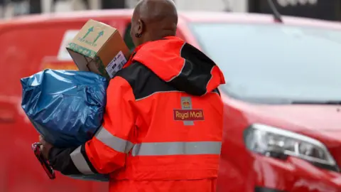 A Royal Mail postman holding two large parcels. He is standing in front of a red Royal Mail van