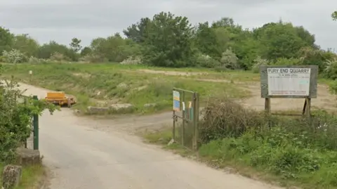 Google Entry to quarry showing road in past green metal gates, a sign saying "Pury End Quarry" on a board to the right and a green area behind the sign with a track through it - part of the quarried land which has been restored