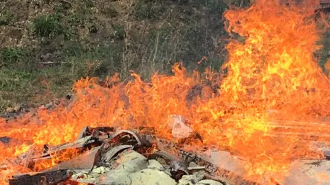 A pile of waste is covered in large bright orange flames. There are green trees in the background behind it.