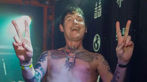Mick Steff Derry Brownson of EMF smiles as he holds both hands in peace signs while shirtless at a performance