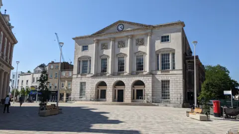 A distant picture of Shire Hall in a sunny day with an analogue clock at the top. It has three arches at ground level, and four columns on the front of the building covering the first and second floors.