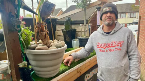  A man is a black woolly hat and grey hoodie stands beside an artificial pond. He has a grey beard and rests his arm on a planks of wood. There is a large plant pot full of stones and an exotic plant. Other plants and a waterfall can be seen. A shed and the wall of a house can been seen close in the background.