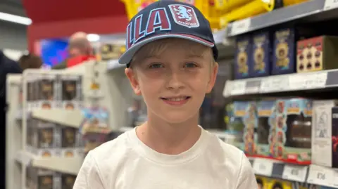 A young boy smiling standing in front of shop shelves, he is wearing a white t-shirt and an Aston Villa baseball cap in the club colours of claret and blue.