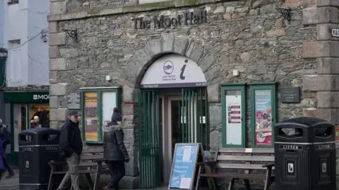 The Lake District National Park Authority tourist office at Moot Hall. It is on the ground floor of the sandstone building and has a green gate in front of the door. Two people are walking into the building. Green noticeboards flank the central doorway, each containing posters. There are two large black litter bins either side and wooden benches in front of the building.