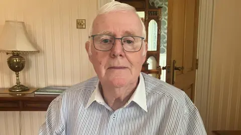 An older man looks into the camera. He has white hair and is wearing a black and white striped shirt with a white collar. He is wearing large square glasses.
