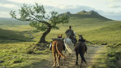 HBO Publicity shot from A Knight of the Seven Kingdoms showing Ser Duncan the Tall riding a white horse with two horses either side of him. He is riding on a path with a curved tree beside him looking towards mountains.