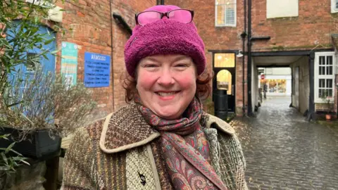 A woman with ginger hair, a pink hat and a patterned coat and scarf standing in a cobbled street with a brick archway behind her.