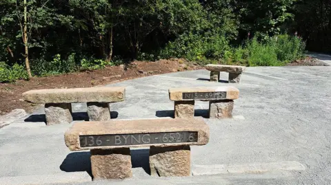 Stone benches which have metal plaques with different carvings are position on a walking trail in Carlisle. The benches, which are place in front of woodland, have a Roman-era design.