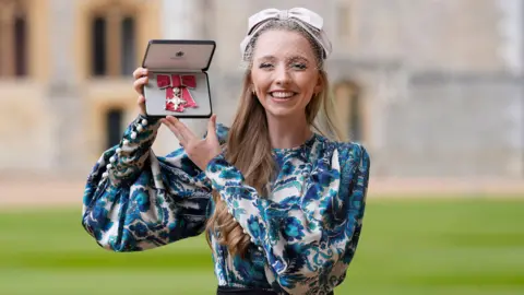 Anna Lapwood at an investiture at Windsor Castle. She is holding her MBE medal which is red and silver. Lapwood is wearing a bow-style fascinator head dress in pale grey with netting covering her forehead and eyes. She is also wearing a blue and grey floral patterned dress. She is smiling as she looks at the camera.