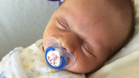 A close-up of an asleep baby sucking on a blue dummy with a fox on it.