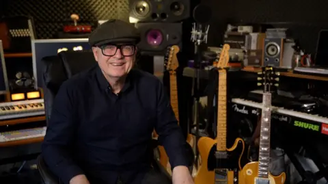 A man dressed all in black, with black glasses and a flat cap. He is sat in a recording studio, smiling at the camera, with three guitars to his left