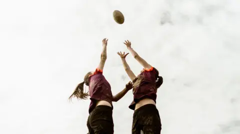 Two girls reaching for a rugby ball in the air. The photo is against a cloudy sky.