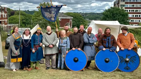 Hrafn Vikings A group of men and women dressed as Vikings, standing in a field with three large circular blue shields in front of them. One member is holding a blue and gold banner on a wooden cross. 