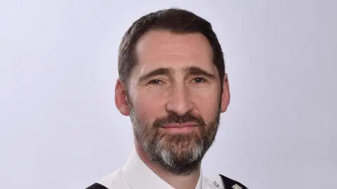 Ben Snuggs, a man with dark hair and a short beard, is pictured facing the camera for a posed professional head shot. His shoulders, which are only just visible, reveal that he is wearing a police uniform. 