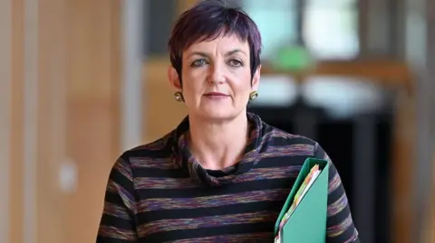 A woman with short purple hair, wearing a purple stripy top and carrying a green folder and black phone in her left hand, looks ahead as she walks down a corridor in the Scottish Parliament 