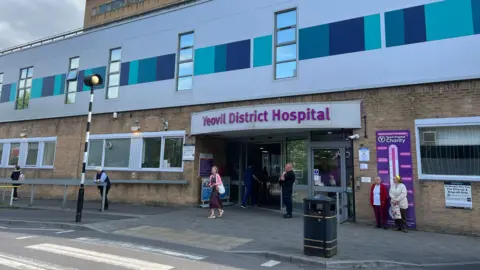 a zebra crossing leading to the main entrance of yeovil district hospital with the hospital's name in purple lettering above an automatic door with a security guard outside. The rest of the hospital building is yellow brick or clad in grey and turquoise panels