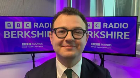 Chris McHugh/BBC Jack Rankin MP smiles for the camera in front of purple BBC Radio Berkshire branding. He is in his early thirties, with short black hair and black rimmed glasses, wearing a dark suit and tie. 