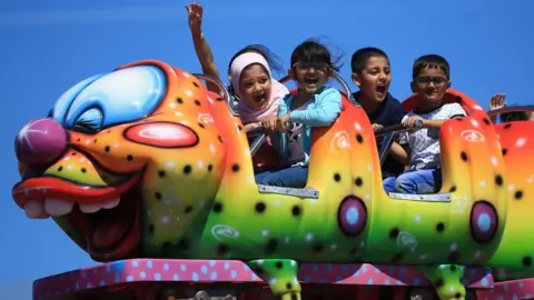 Getty Images Two girls and two boys sit and scream in a large caterpillar fairground ride on a sunny day.