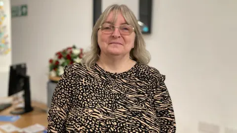 Woman in her mid-50s standing in front of a desk in an office smiling into a camera. She is wearing a black and gold patterned dress with light grey hair and glasses. 