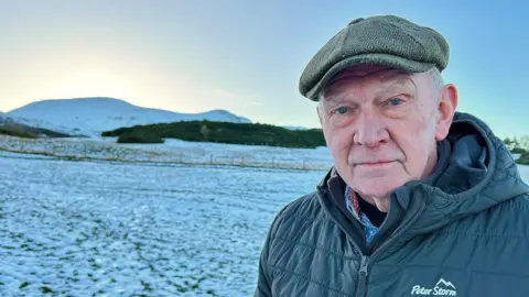 BBC Tom Wood is in the snowy Pentland hills. This is his head and shoulders shot. He is wearing a green cap and a thick winter jacket. 