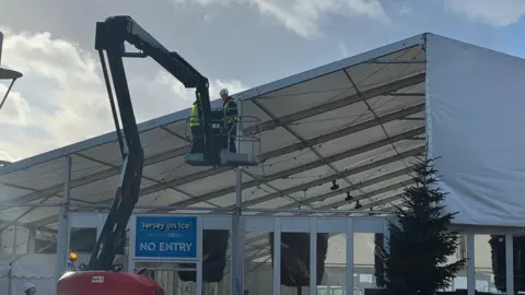 A large white marquee tent is checked on outdoors under a partly cloudy sky. Two workers wearing safety gear are elevated on a hydraulic boom lift, working near the top edge of the tent structure.