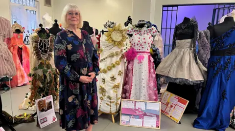 Marjorie Wells stands next to her dresses - one is white and decorated with gold flowers and has a sunflower on top. The other is white and decorated with pink flowers.