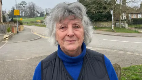 John Devine/BBC Elaine Wakerley, 73, has short grey hair and is wearing a blue polo neck jumper, with a black body warmer on top. Behind her is a rural road going up a hill, with a grassy field with trees visible behind her. There are properties either side of the road.