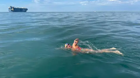 Melanie Barratt/FB A picture of Melanie Barratt swimming in the blue sea in a colourful swimsuit and cap. There is a ship in the background.